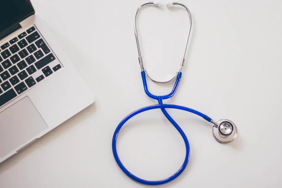 A blue stethoscope lying next to a laptop on a white desk